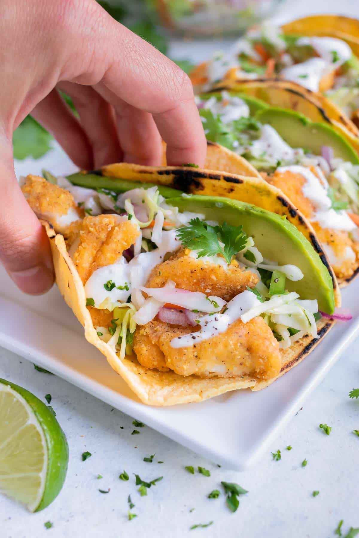 A hand is used to lift a fish taco with lime crema from the plate.