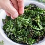 Air fryer kale chips are lifted out of a white bowl from the counter.