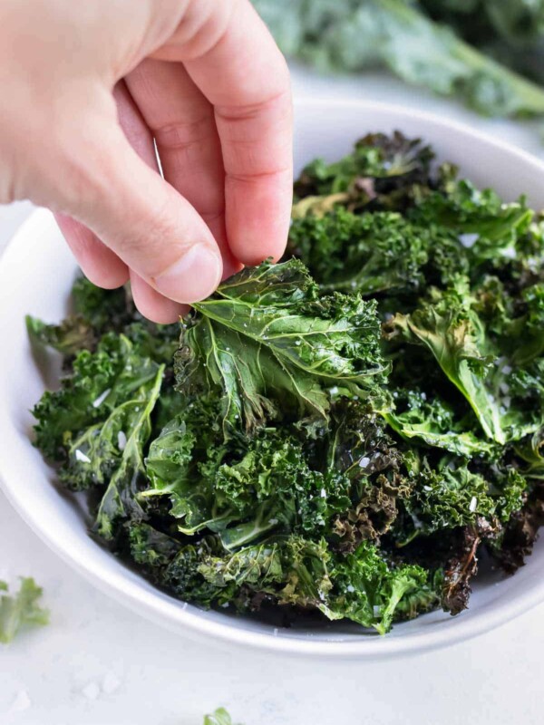 Air fryer kale chips are lifted out of a white bowl from the counter.