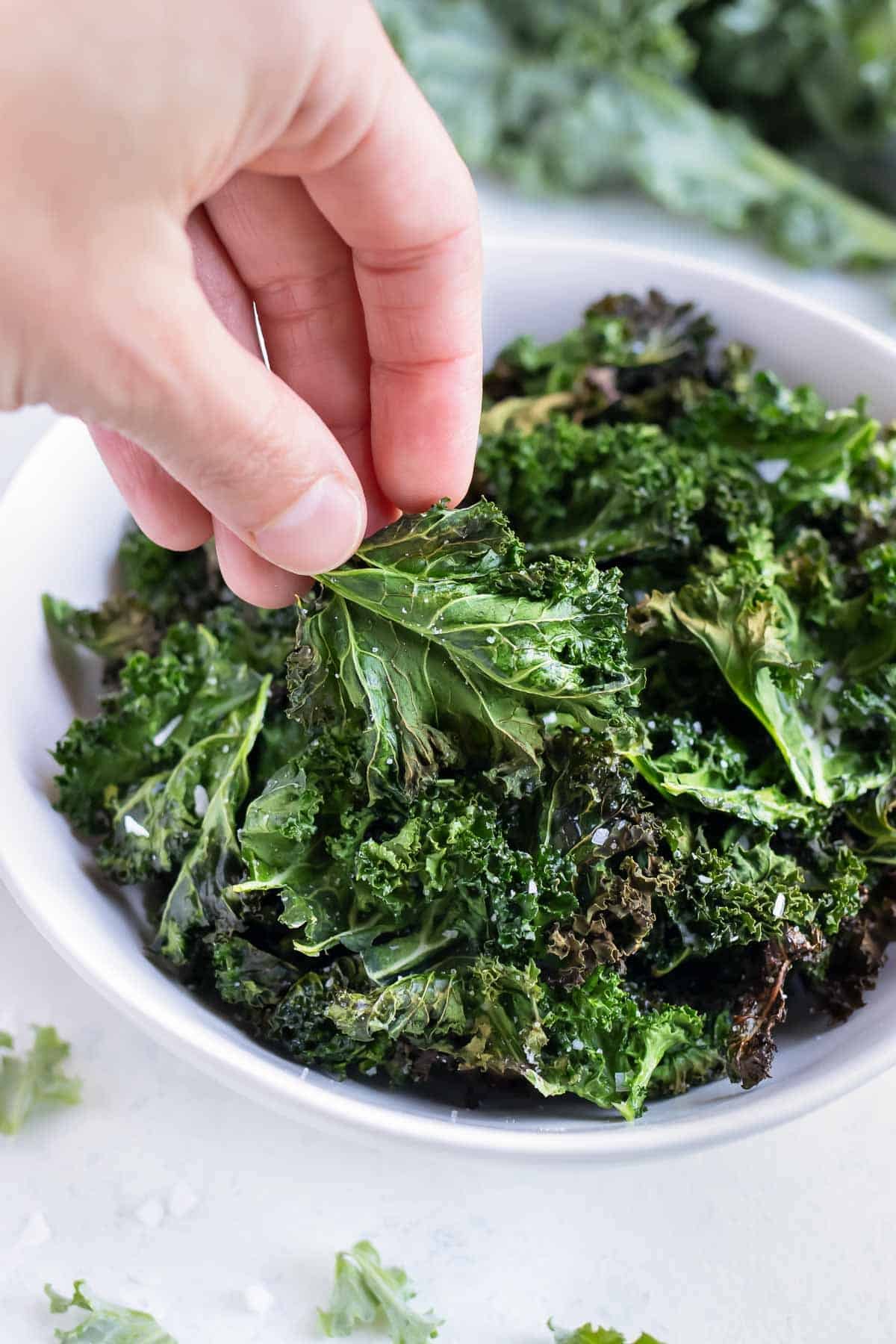 Air fryer kale chips are lifted out of a white bowl from the counter.