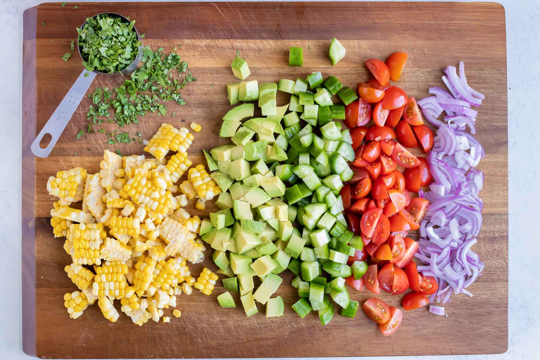 Corn, avocado, red onion, tomato, cilantro, and cucumber are chopped on a cutting board.