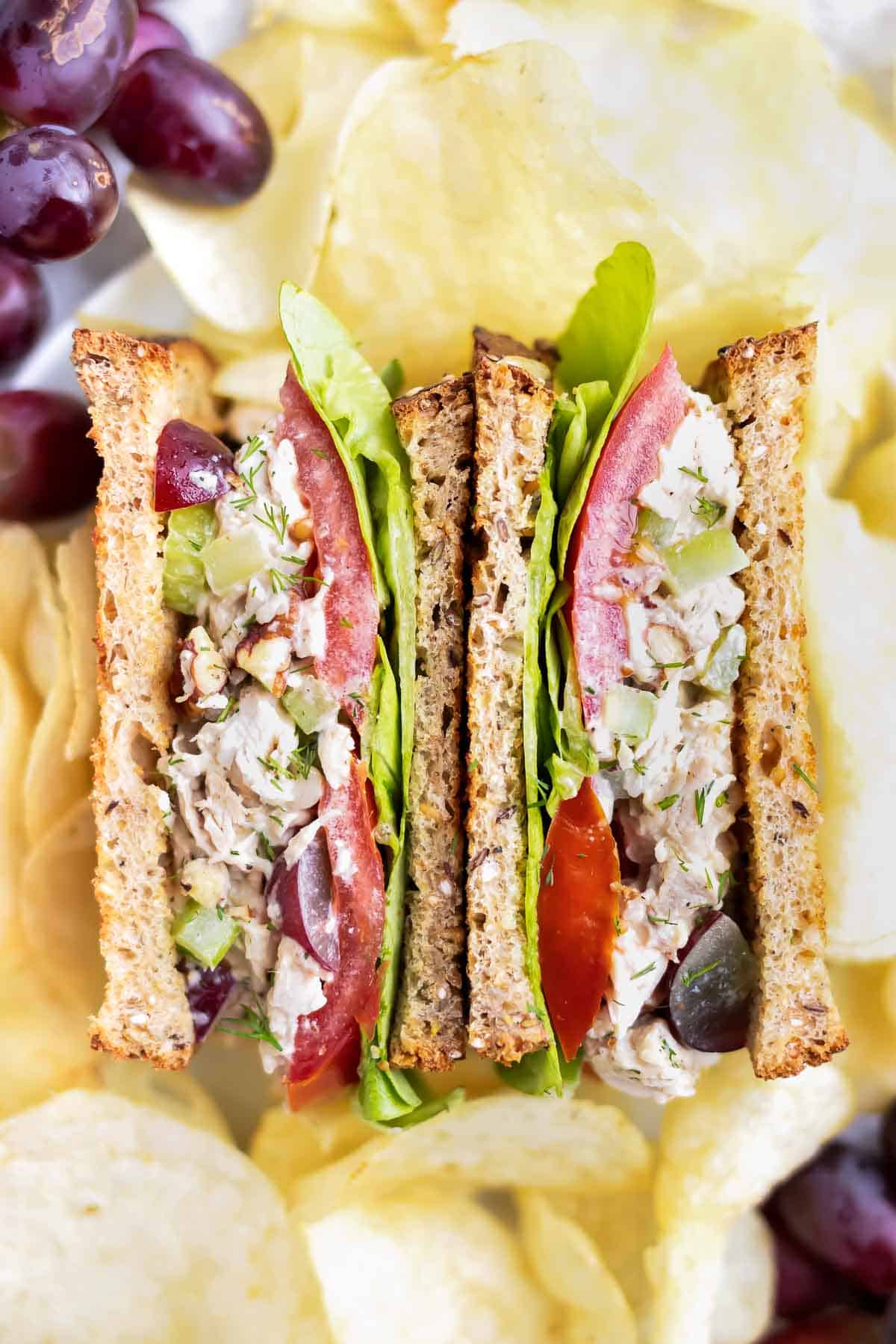 Two halves of a chicken salad sandwich surrounded by potato chips and red seedless grapes.