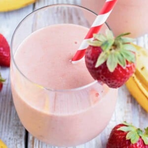 A glass full of a strawberry and banana smoothie with a strawberry as a garnish and a red/white striped straw.