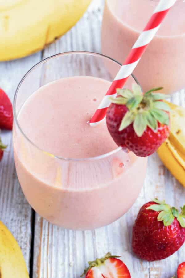 A glass full of a strawberry and banana smoothie with a strawberry as a garnish and a red/white striped straw.