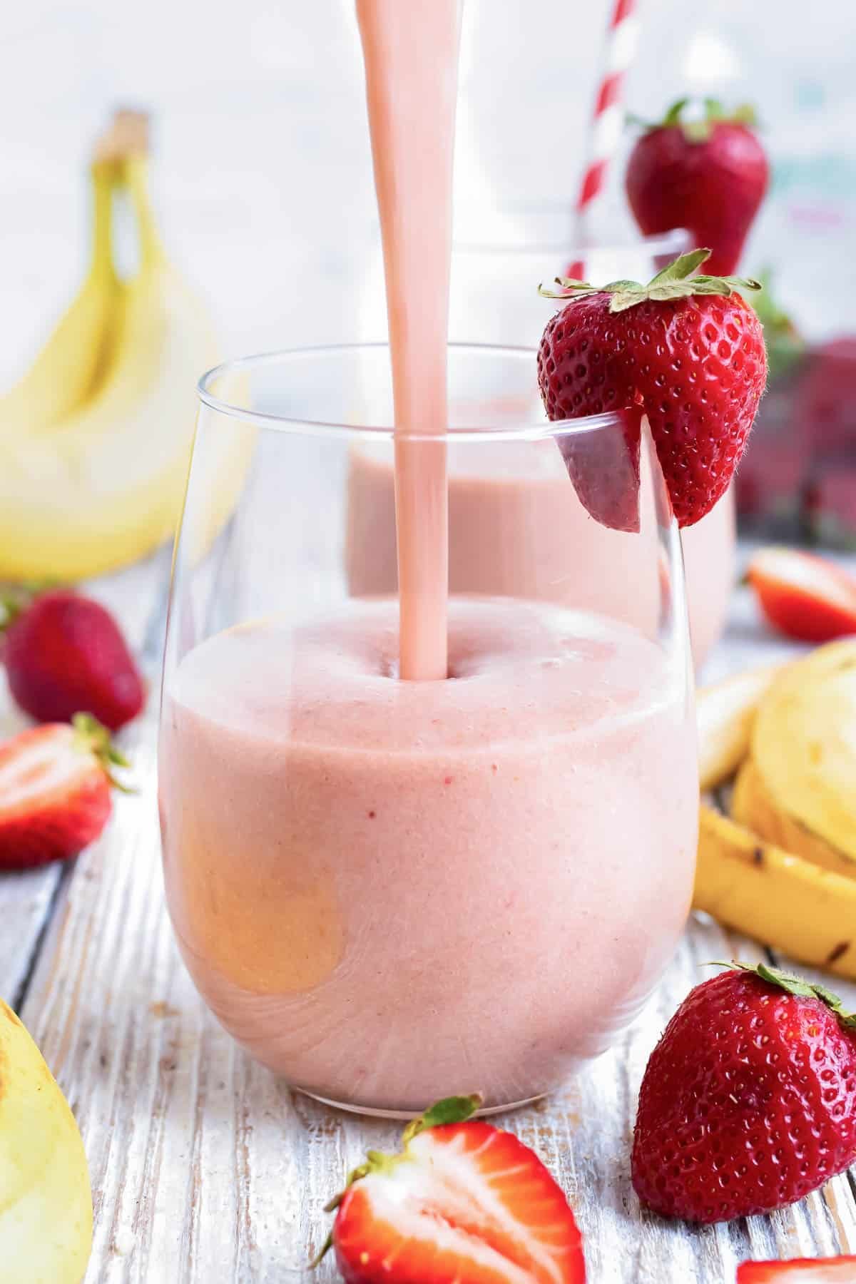 Strawberry banana smoothie being poured into a glass with strawberries and bananas around it.