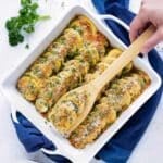 A hand picking up a serving of a healthy squash casserole recipe from a white porcelain baking dish and a blue towel underneath it.