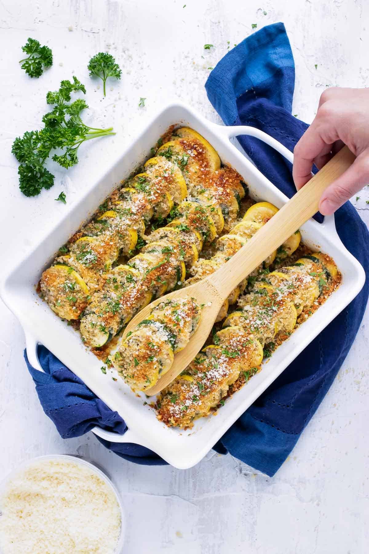 A hand picking up a serving of a healthy squash casserole recipe from a white porcelain baking dish and a blue towel underneath it.