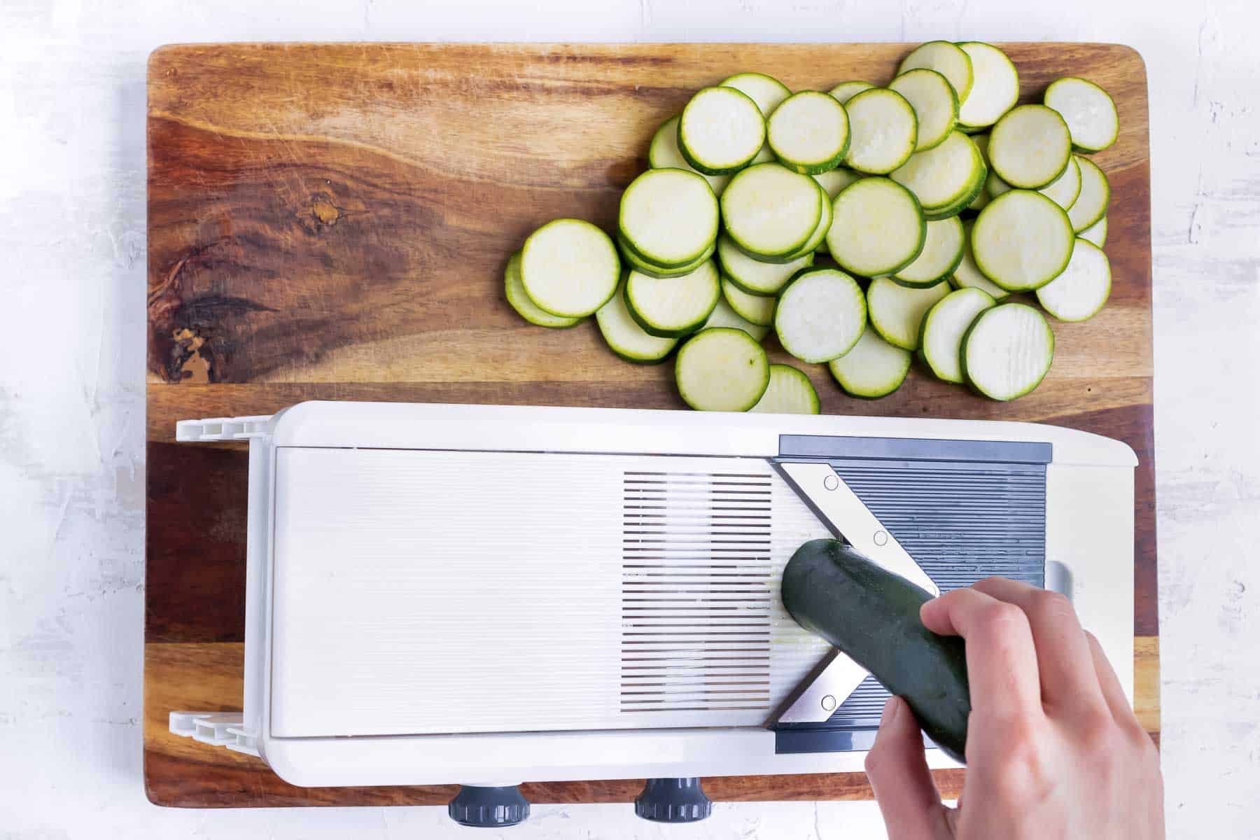 A hand using a mandolin to thinly slice zucchini squash for a squash casserole.