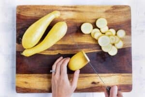 A hand carefully slices a yellow squash into thin slices for a summer casserole recipe.
