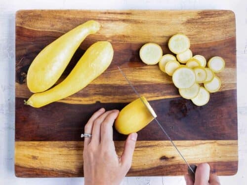 A hand carefully slices a yellow squash into thin slices for a summer casserole recipe.