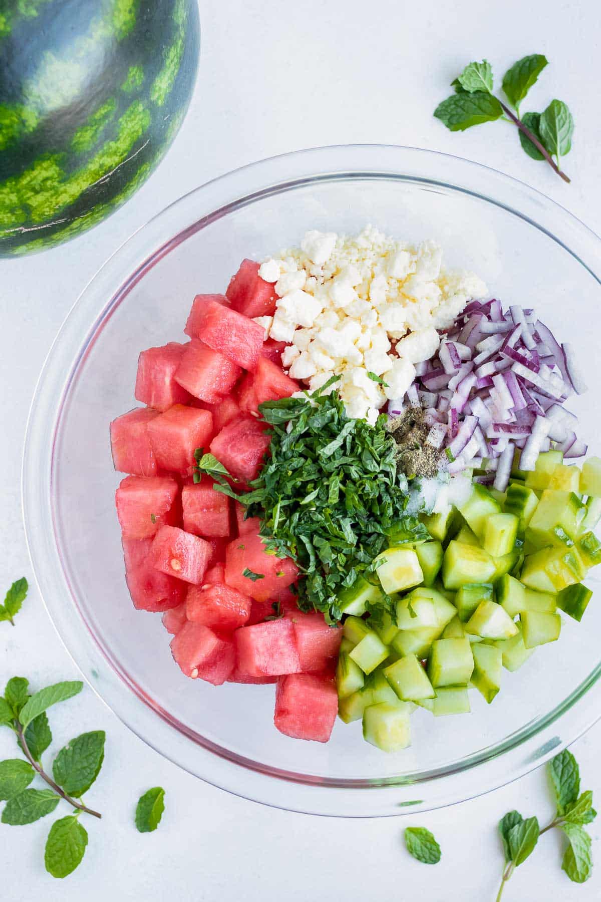 A clear glass mixing bowl full of cubed watermelon, feta cheese crumbles, red onion, cucumbers, and chopped sweet mint.