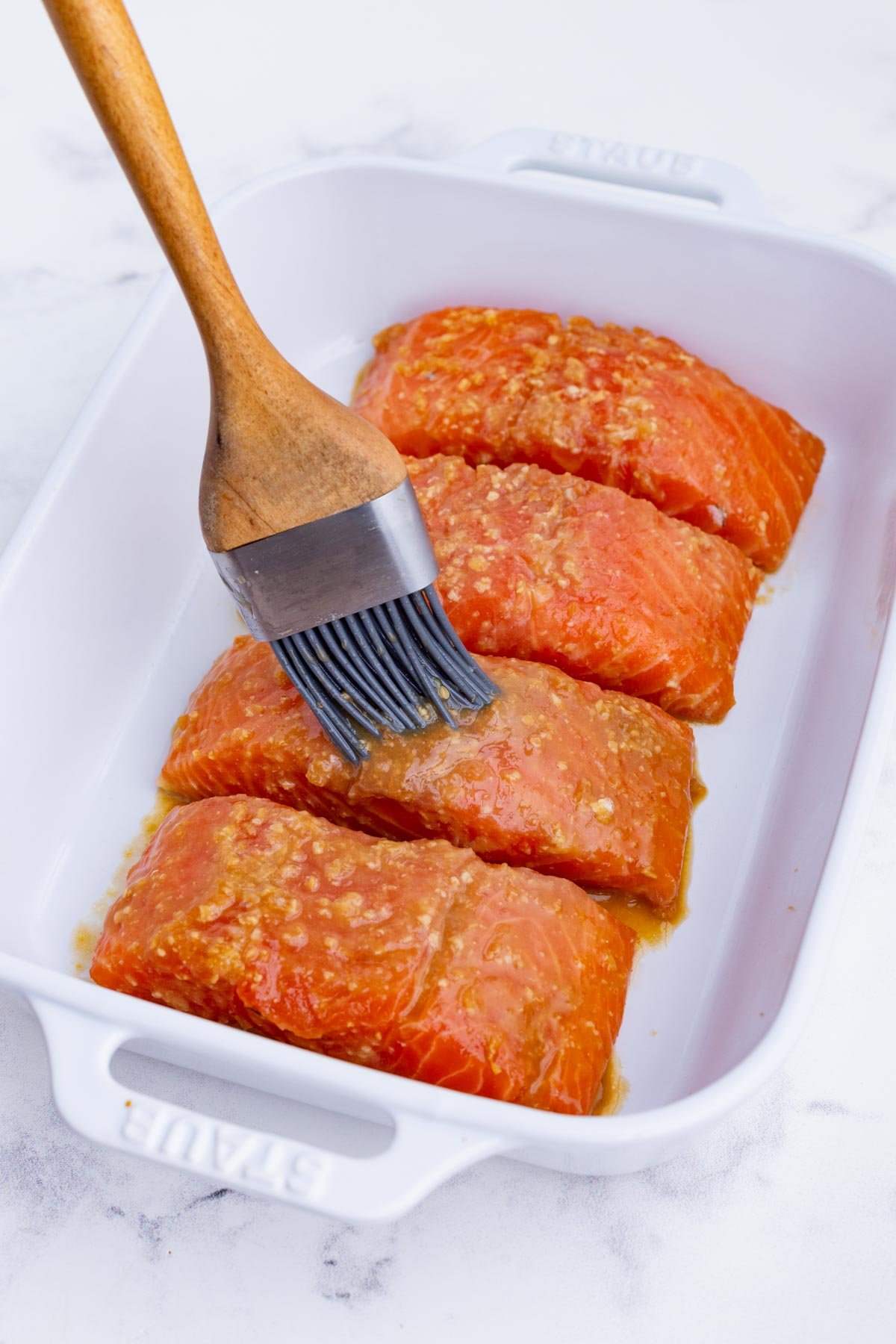 Sauce is brushed on top of salmon in a white baking dish.
