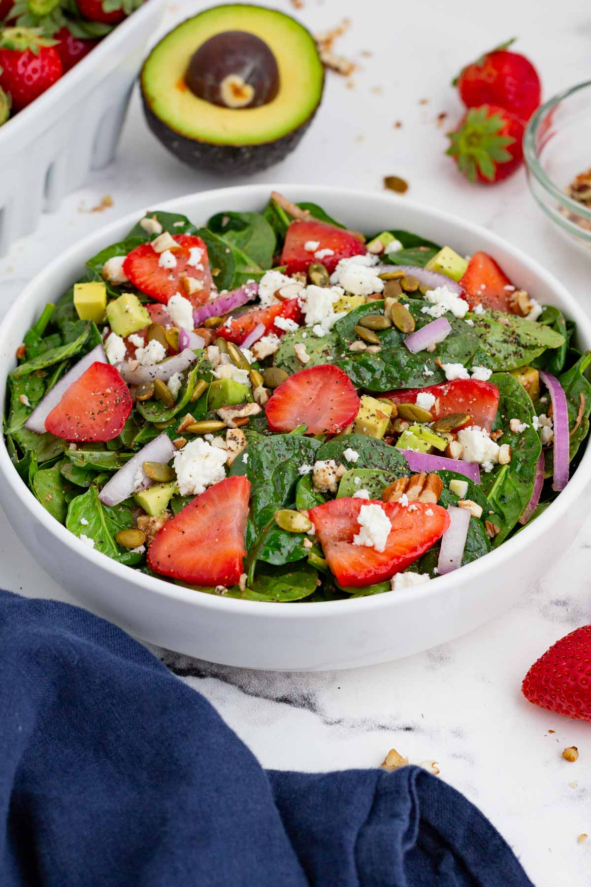 A strawberry spinach salad in a white bowl with an avocado behind it.