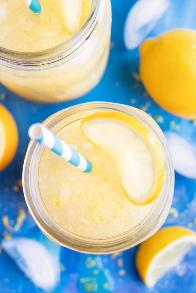 Two glasses of homemade frozen lemonade are served with a straw next to ice cubes.