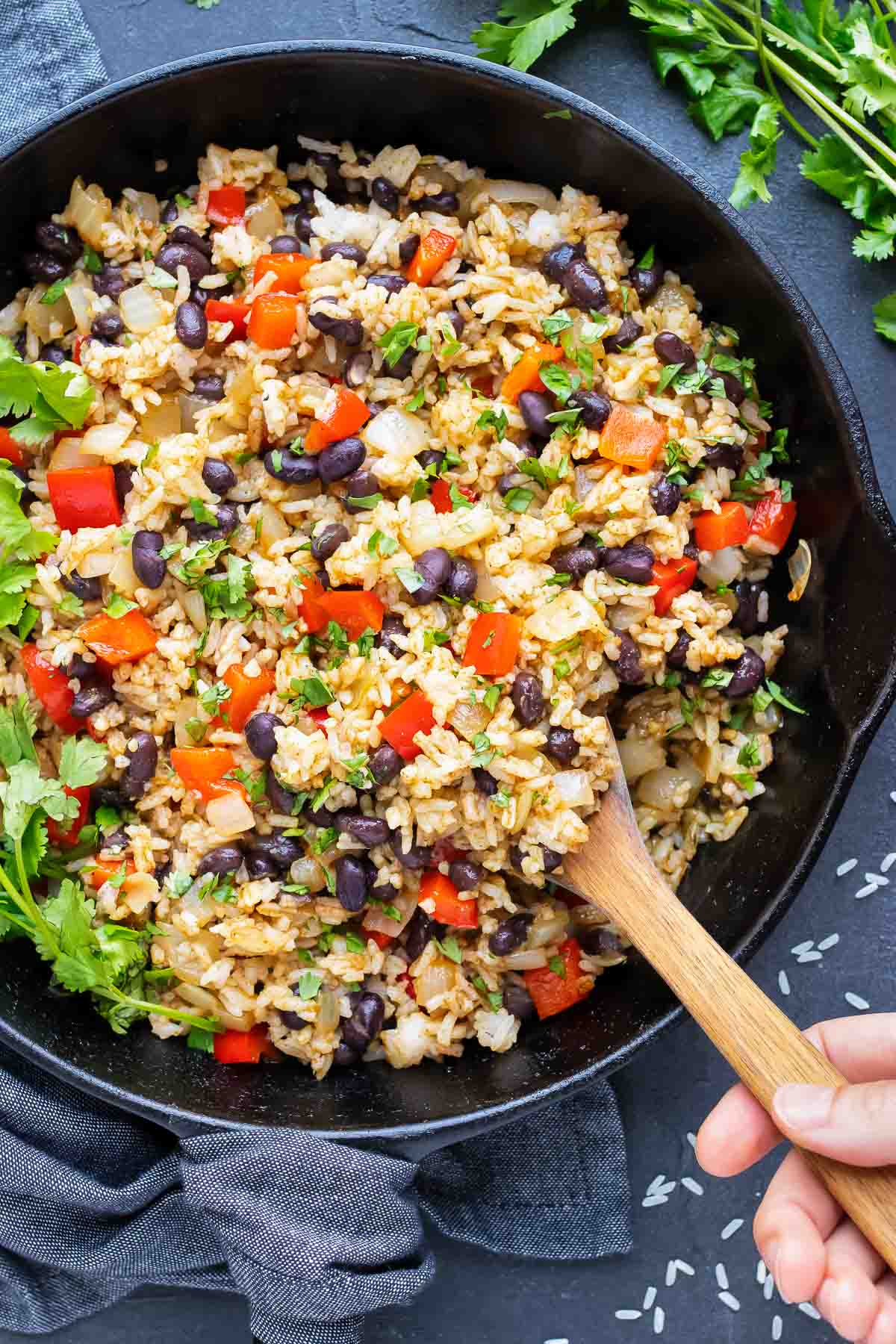 A hand scooping out some gallo pinto from a cast-iron skillet with a wooden spoon.