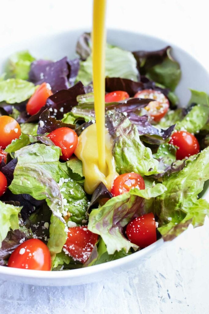 Pouring 5-minute honey mustard vinaigrette over a salad with mixed greens and cherry tomatoes.