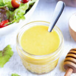 A jar full of honey mustard dressing with a silver spoon next to two wooden salad bowls.