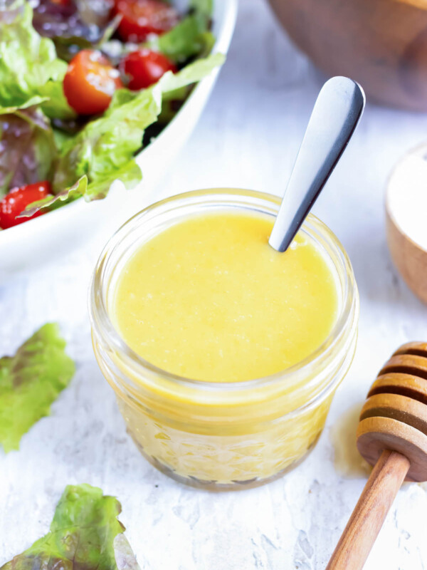 A jar full of honey mustard dressing with a silver spoon next to two wooden salad bowls.