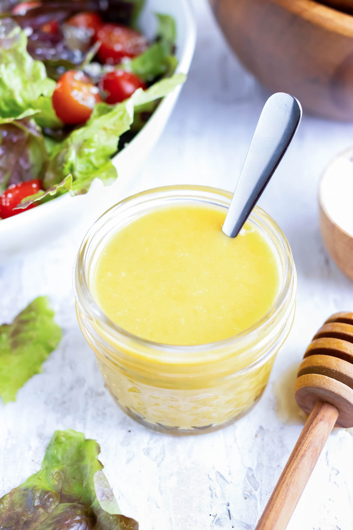 A jar full of honey mustard dressing with a silver spoon next to two wooden salad bowls.