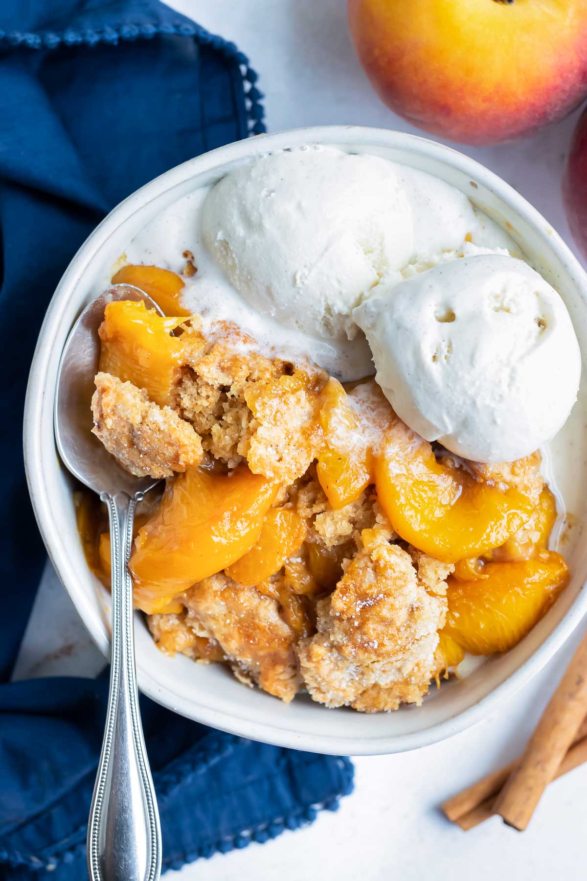 A metal spoon is picking up sweet, warm peaches and flakey cobbler crust of a homemade peach cobbler topped with ice-cream in a bowl.