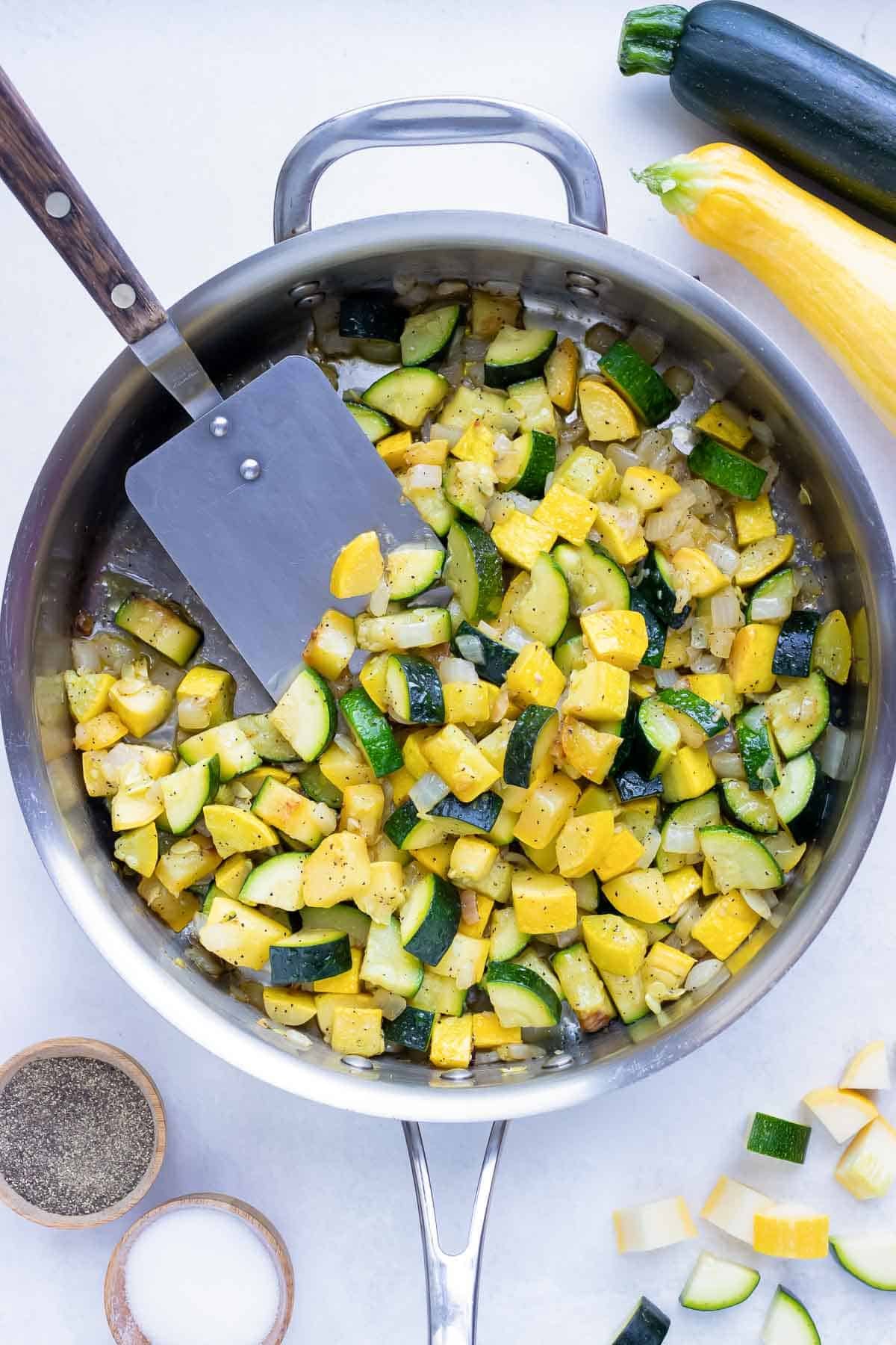 Sautéed zucchini and yellow summer squash in a stainless steel skillet with a spatula.