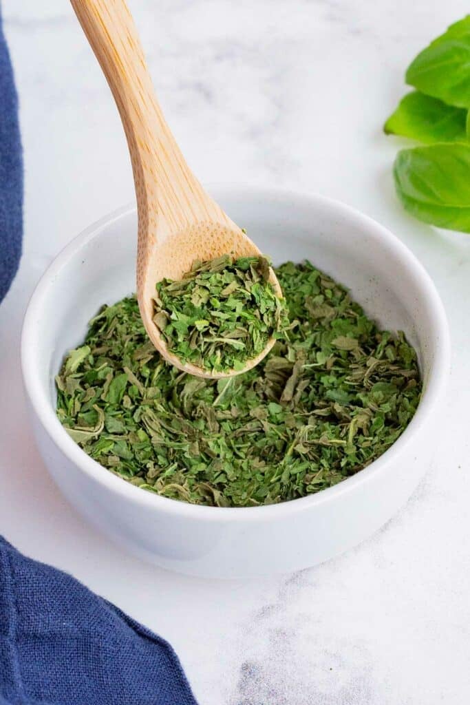 Dry, crushed up basil in a white, ceramic bowl with a wooden spoon.