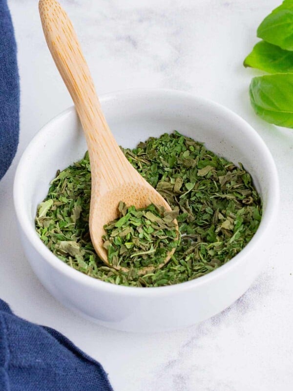 Dry, crushed up basil in a white, glass bowl with a wooden spoon.