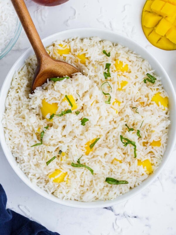A wooden spoon scoops up some mango coconut rice with coconut milk from a white serving bowl.