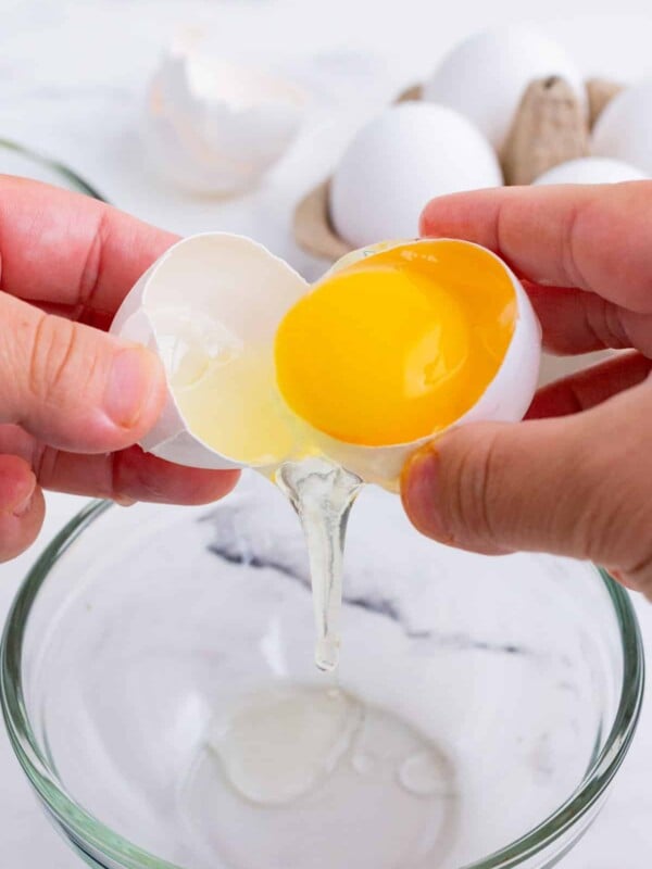 A cracked open egg with an egg yolk being tossed between the shell halves with the whites falling into a bowl beneath.