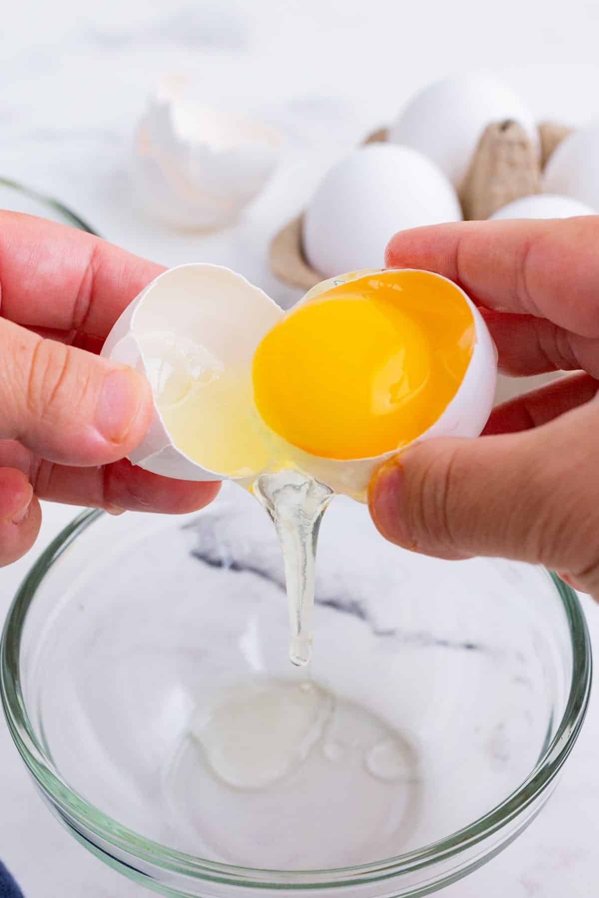 A cracked open egg with an egg yolk being tossed between the shell halves with the whites falling into a bowl beneath.