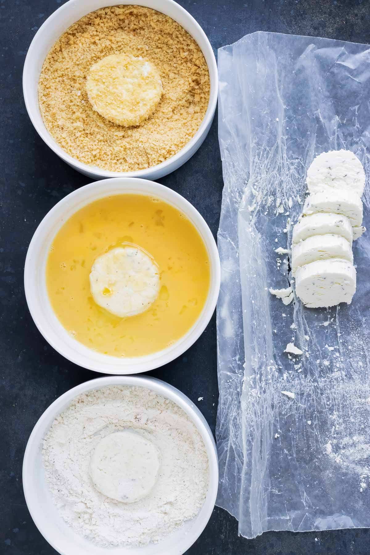 Three bowls of breadcrumbs, flour, and eggs next to goat cheese medallions showing how to make fried goat cheese.