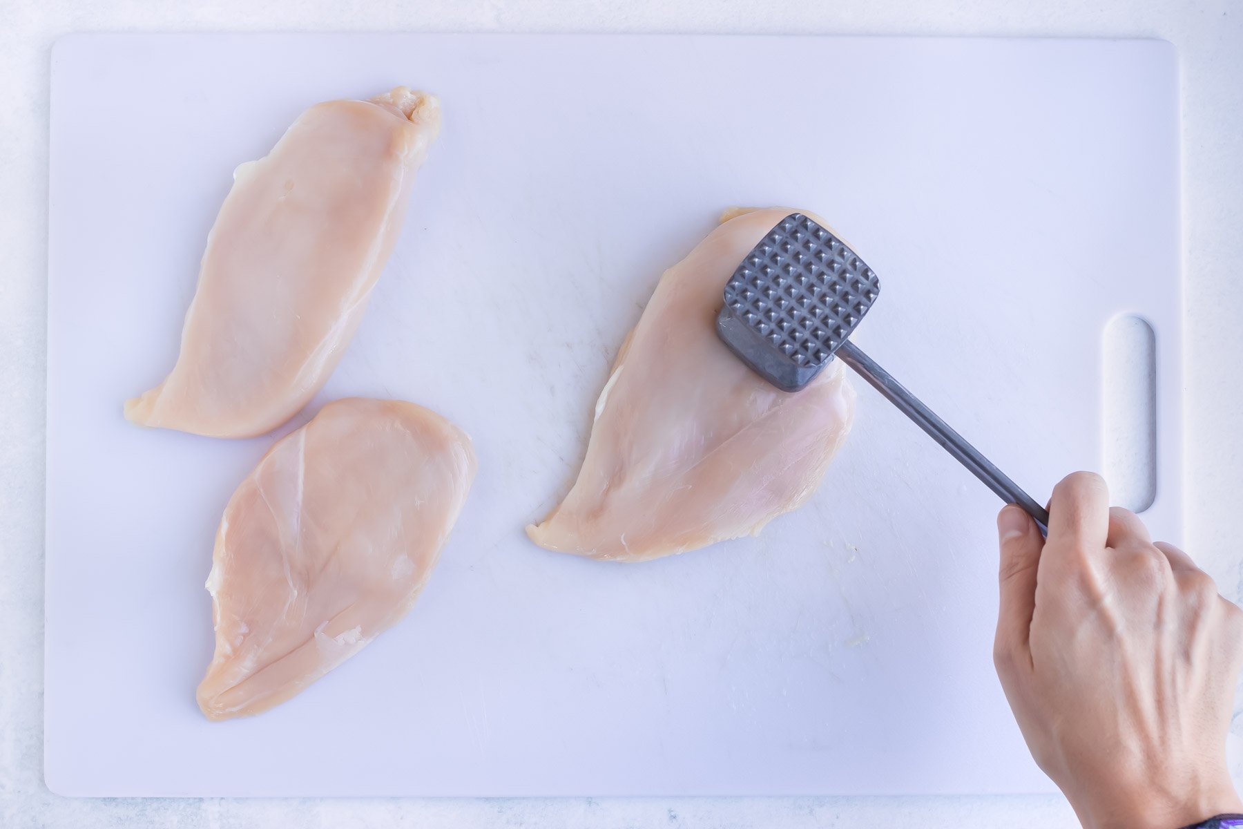 A hand hitting a piece of chicken to show how to tenderize with a meat mallet.