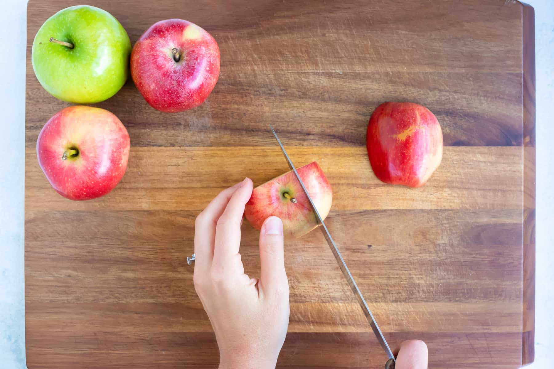 Cutting the side of an apple with a knife  on a cutting board with other apples waiting to be cut.