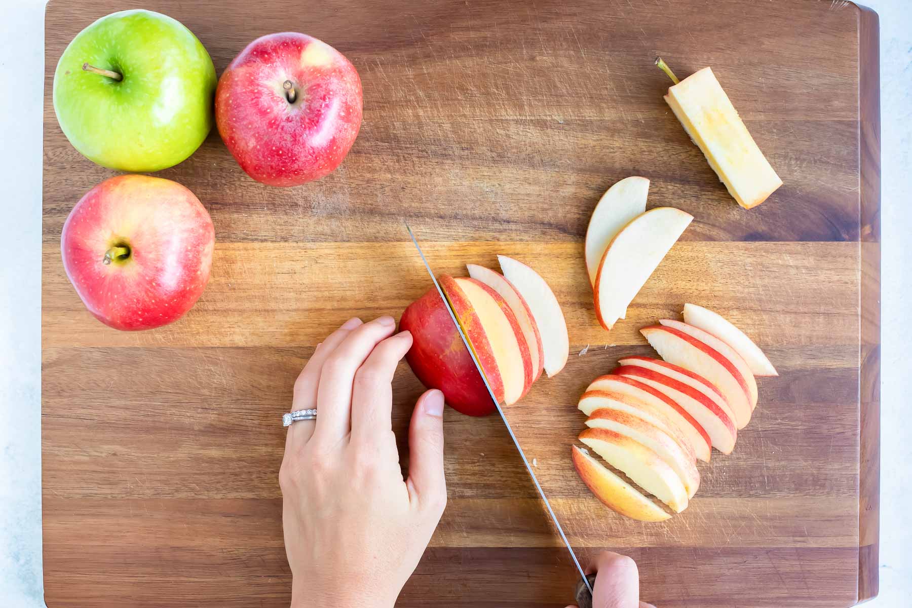 Cutting apple slices with a knife on a cutting board with other apples waiting to be cut and the core to the side.