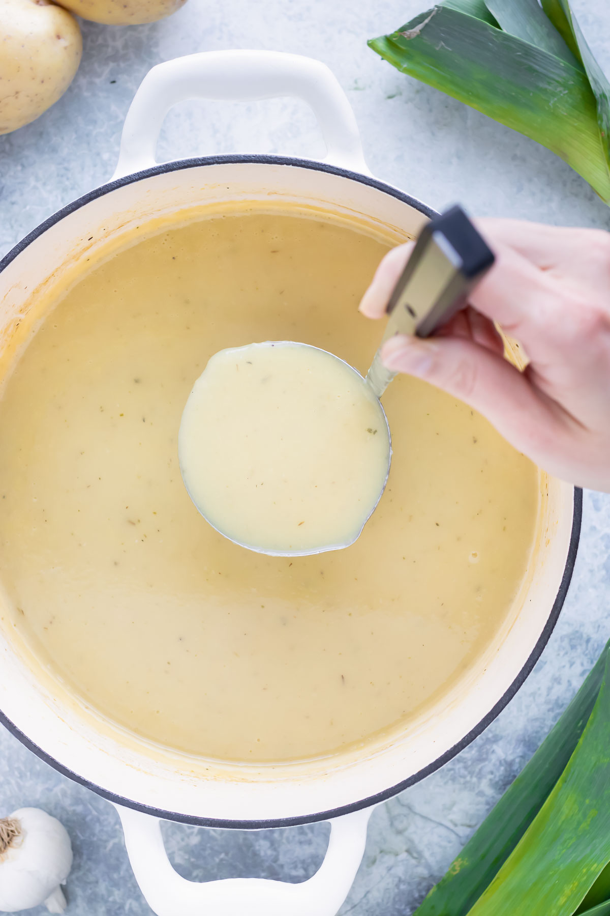 A soup ladle scooping out a serving of a healthy and creamy vegan potato leek soup from a Dutch oven.