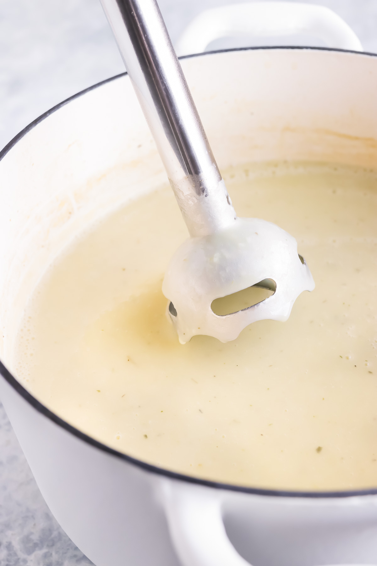 An immersion blender coming out of a large pot full of a creamy potato soup that was just blended up.