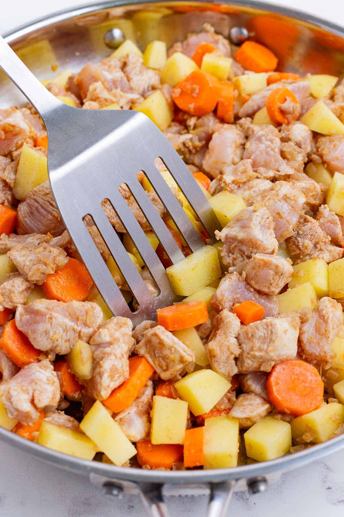 A spatula stirs the chicken, carrots, and potatoes into onions.