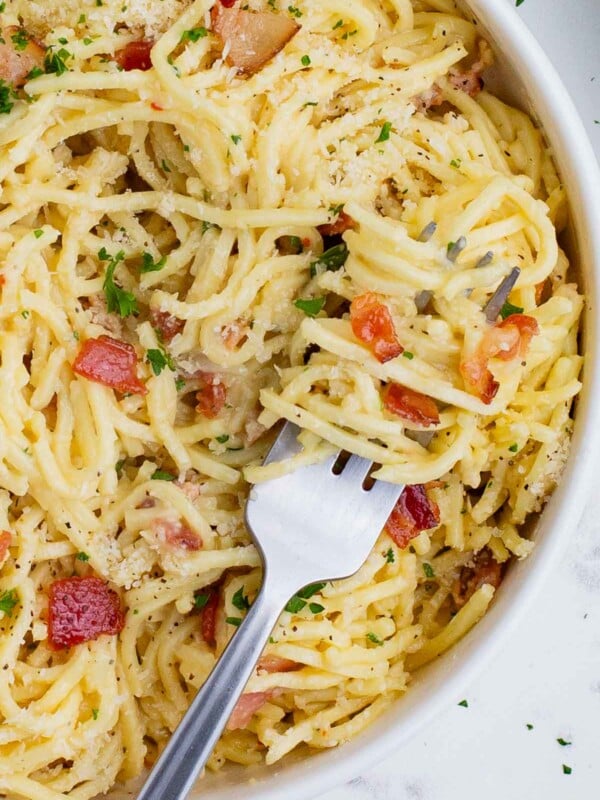 A fork digs into a big bowl of pasta carbonara filled with bacon, cheese, and fresh herbs.