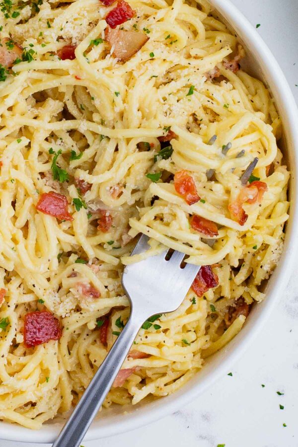 A fork digs into a big bowl of pasta carbonara filled with bacon, cheese, and fresh herbs.