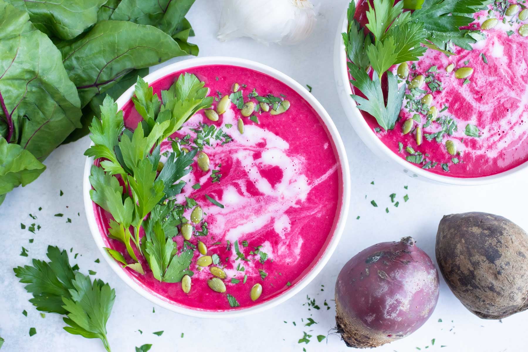 Two bowls of creamy beet soup with fresh beets and parsley on a white counter.