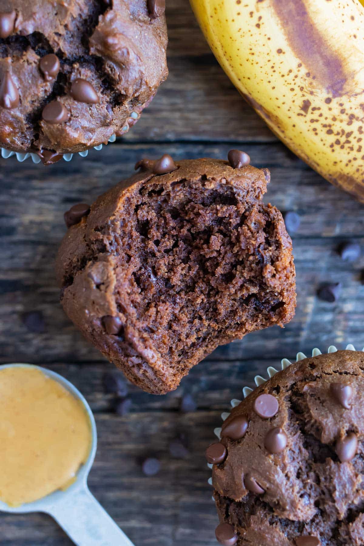 A chocolate muffin with a bite taken out of it, is laid on the counter next to ingredients from this recipe.