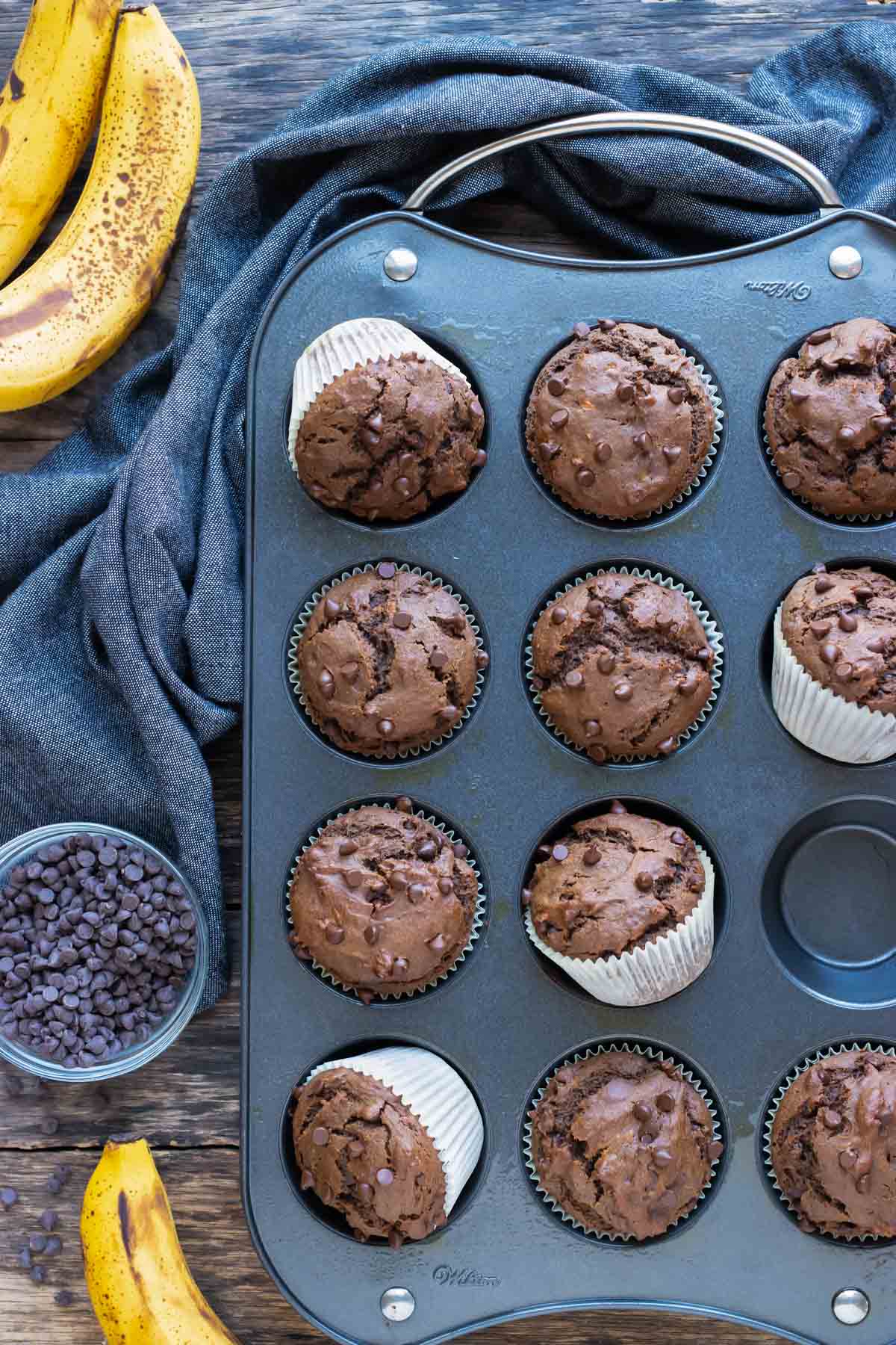 Healthy chocolate banana muffins in a muffin pan with bananas and chocolate chips around them.