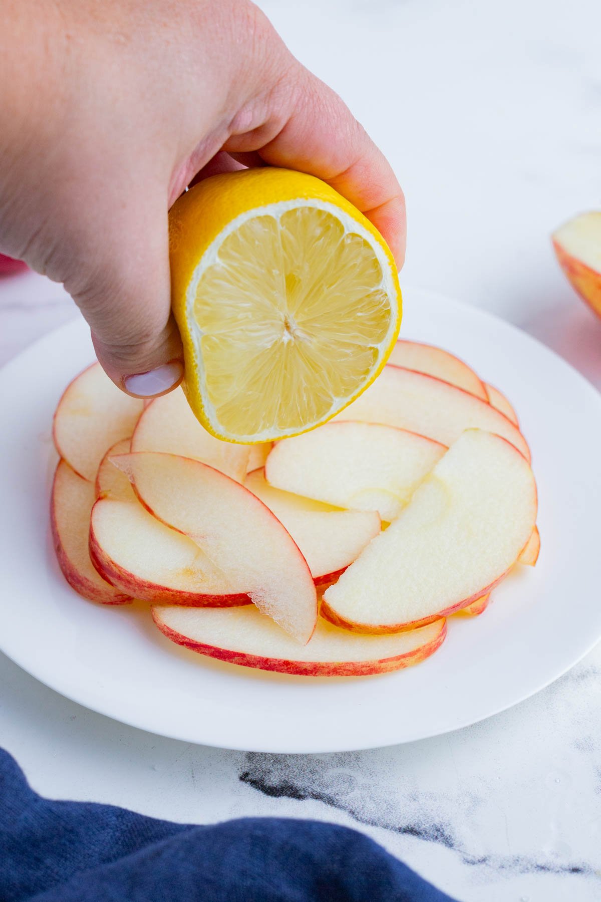 Lemon being squeezed over thinly sliced apples on a white plate.
