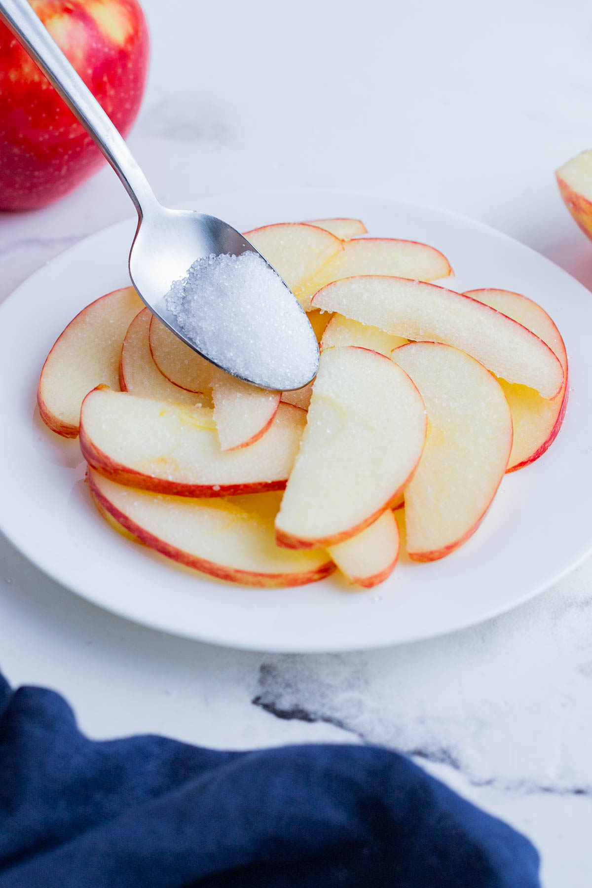 Citric acid being sprinkled with a spoon over thinly sliced apples on a white plate.