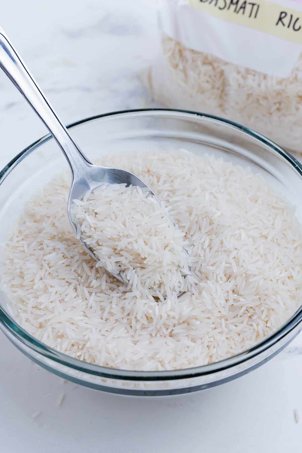 A spoon scoops Basmati rice out of a glass bowl.