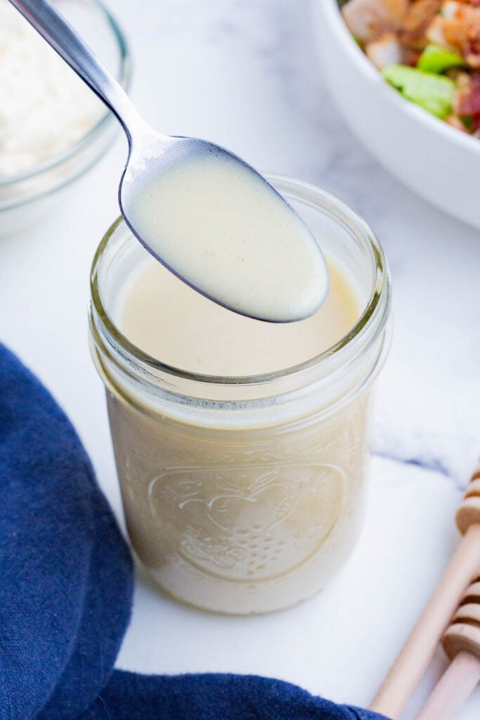 A ﻿spoon scoops up a serving of a creamy cobb salad dressing made from oil, honey, mayo, and mustard from a glass jar.