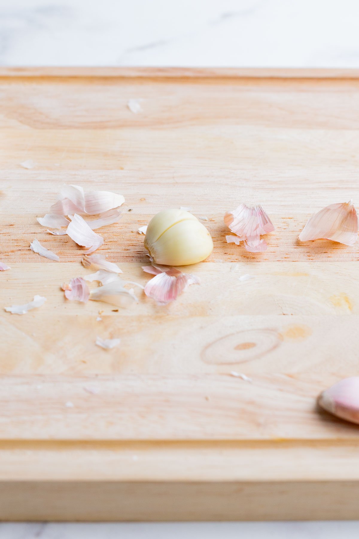 A lone peeled garlic clove surrounded by its outside papery skin on a cutting board.