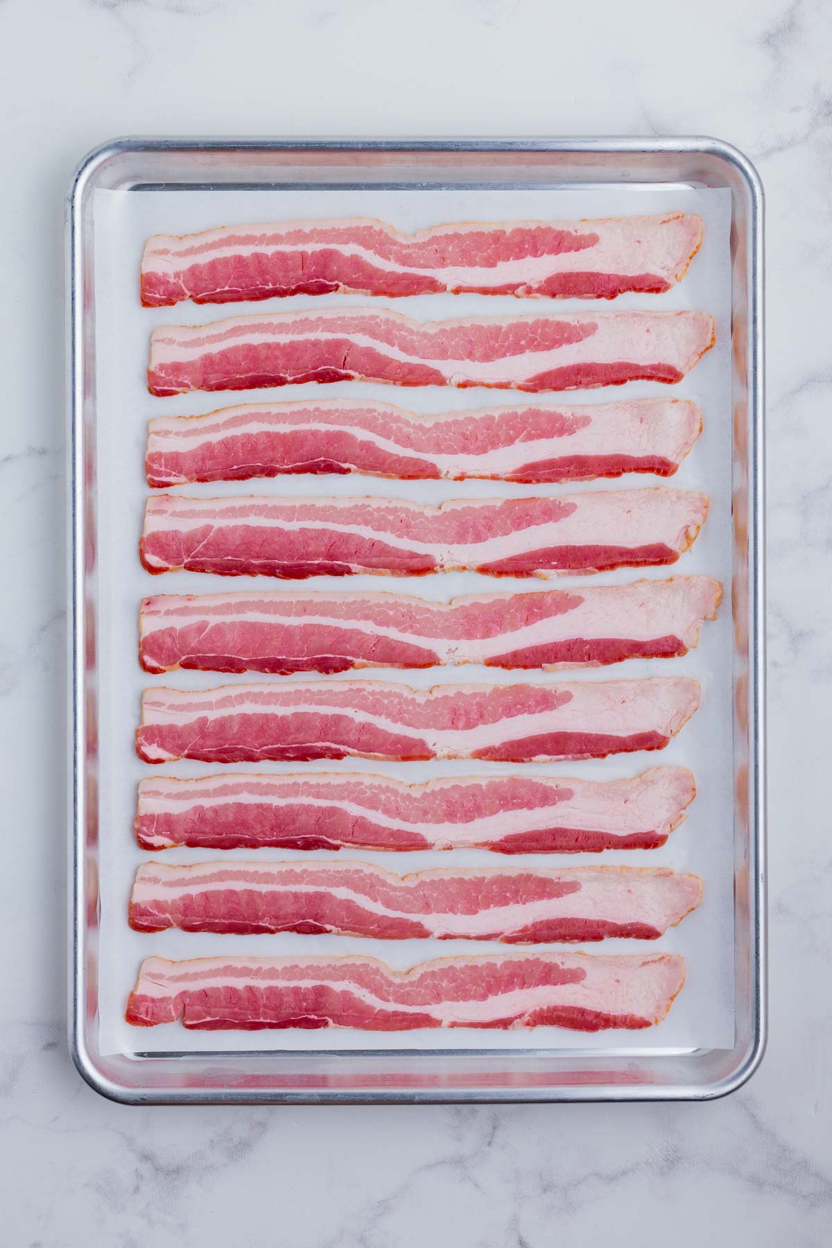 Raw bacon is lined up on a baking sheet lined with parchment paper.