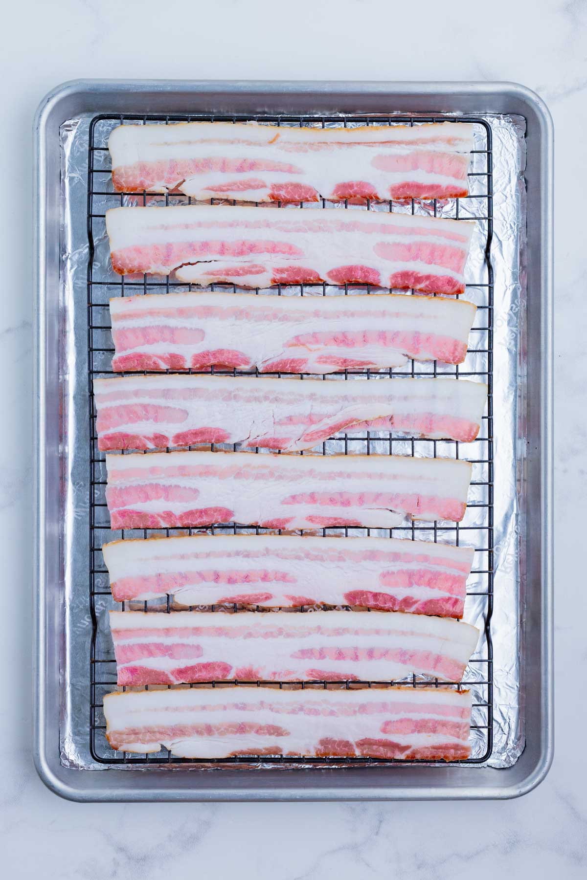 Raw bacon is lined up on a baking sheet with a wire rack.