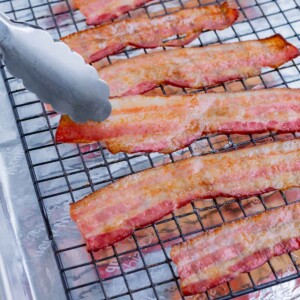 Bacon cooked on a wire rack in a baking sheet is picked up with tongs.